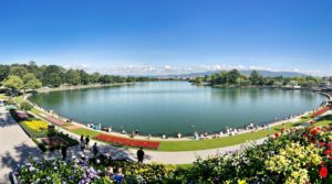 A wide-angle view of a serene lakeside park in Geneva, featuring vibrant greenery, blooming flowers, and people picnicking and strolling by the water under a clear blue sky, showcasing the tranquility of parks gardens Geneva.