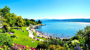 A scenic view of Lake Geneva, featuring families enjoying leisurely walks along the water's edge, children playing on the grass, and picnicking under trees, surrounded by vibrant green foliage and the shimmering blue lake, under a clear blue sky, perfect for