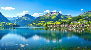 Panoramic view of serene alpine pastures near Geneva, featuring a tranquil lake surrounded by lush green mountains, a charming medieval village in the hills, and clear blue skies, ideal for showcasing the peaceful atmosphere of alpine pasture hikes near Geneva.