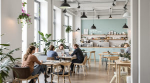 A stylish, minimalistic coworking space in Geneva with young professionals working on laptops. The light grey walls and mint green accents complement the natural wooden furniture. Large windows fill the room with natural light, while potted plants add freshness. In the background