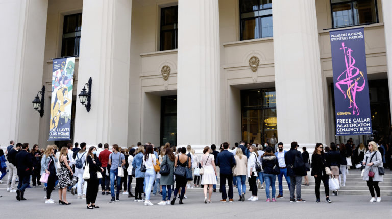 A diverse group of visitors dressed in stylish casual outfits enjoys a free outdoor cultural exhibition outside the Palais des Nations in Geneva. The elegant architecture of the building, with its neutral tones and artistic event banners, sets a calm and sophisticated atmosphere, reflecting a vibrant