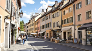A vibrant urban street view in a picturesque Geneva neighborhood, highlighting local shops and markets bathed in natural daylight. The scene features charming architecture with earthy tones and pastel façades, showcasing everyday life and details that reflect the community's spirit, appealing to gene