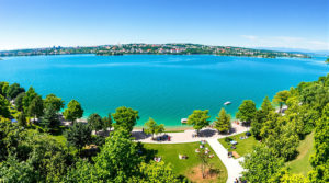 A wide-angle view of a peaceful lakeside park in Geneva, showcasing lush green trees and vibrant blue waters. Groups of people are picnicking, walking, and boating under a clear sky, highlighting the natural beauty and open space, perfect for a