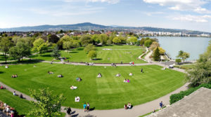 Panoramic view of a vibrant park in Geneva, showcasing lush green lawns and lakeside paths filled with people picnicking, jogging, and relaxing under blooming flowers and gentle hills, capturing the serene beauty of free weekend parks Geneva in soft natural light