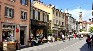 A lively street scene in a charming Geneva neighborhood featuring local shops and a busy market during the day. The buildings showcase earthy tones and pastel façades, highlighting unique architecture. The atmosphere reflects student-friendly living, with bustling streets and peaceful corners, capturing the