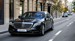 A sleek black 2024 Mercedes S-Class drives smoothly down a serene street in Geneva, with a professional chauffeur in a sharp suit behind the wheel. The car features a 'SWISS LIM CO' white license plate, representing elegance and reliability. The