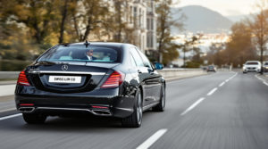 A polished black 2024 Mercedes S-Class drives along a scenic road in Geneva, showcasing a professional chauffeur in a tailored suit through the window. The car, featuring a white license plate 'SWISS LIM CO', is set against a discreet urban background