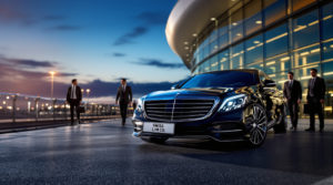 A black 2024 Mercedes S-Class is parked at the Geneva airport terminal, displaying a 'SWISS LIM CO' license plate. Sophisticated chauffeurs in formal attire stand nearby, with a backdrop of deep blue skies, creating an atmosphere of elegance